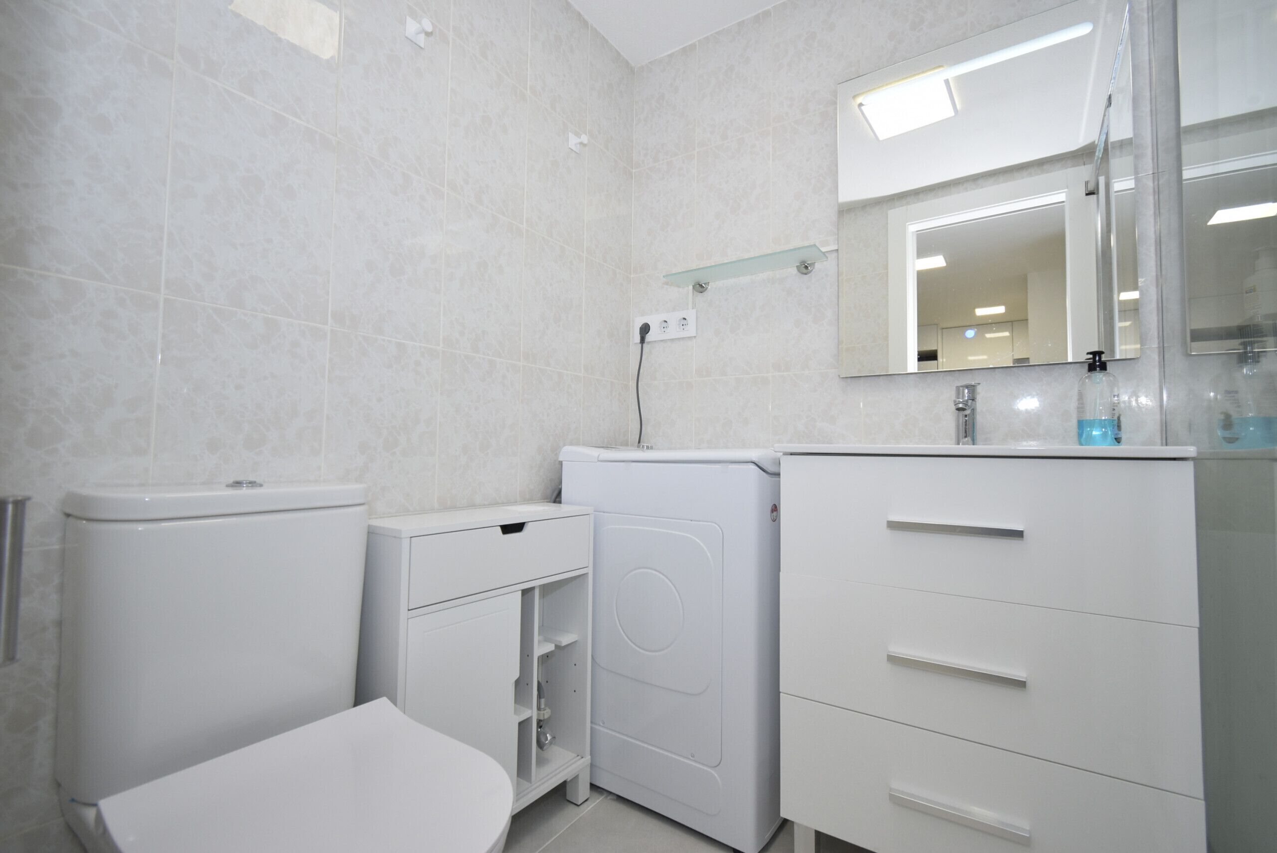 A bathroom with a toilet, washing machine, and a sink with a mirror above it.