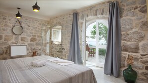 A bedroom with a stone wall, a bed with a striped bedspread, a bedside table, a mirror, and a view of the outdoors through a glass door.