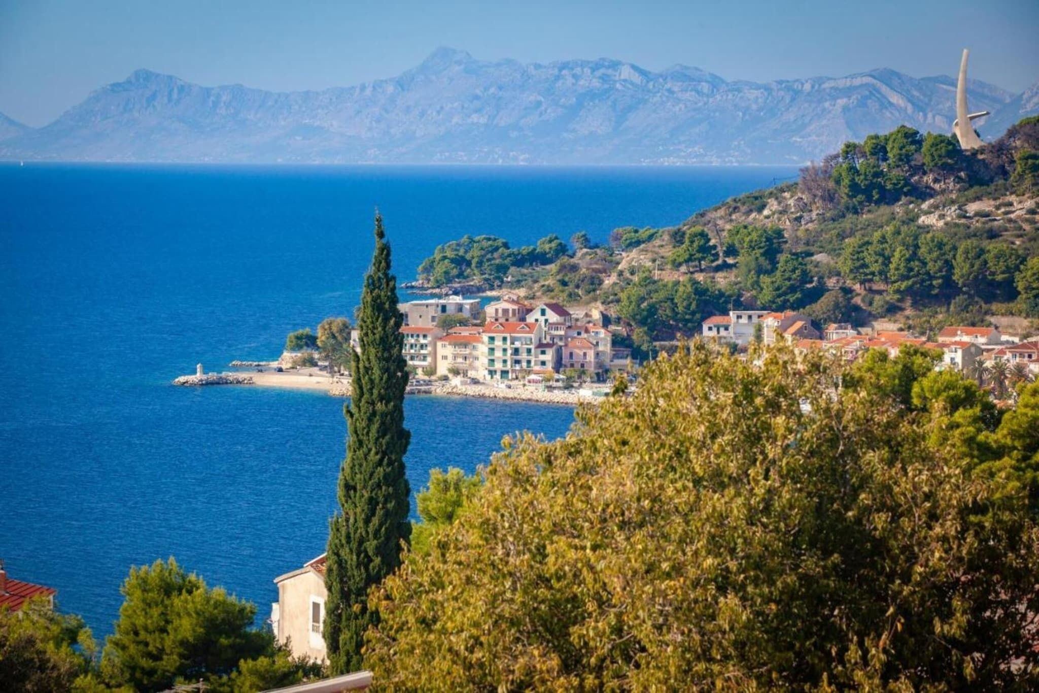 Un poble costaner amb edificis al llarg de la riba, una imponent agulla d'església i una vista clara del mar i de les muntanyes llunyanes.