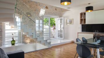 Un intérieur moderne avec un escalier menant à un balcon, un coin repas doté d’une table en verre et une cuisine équipée d’un téléviseur.