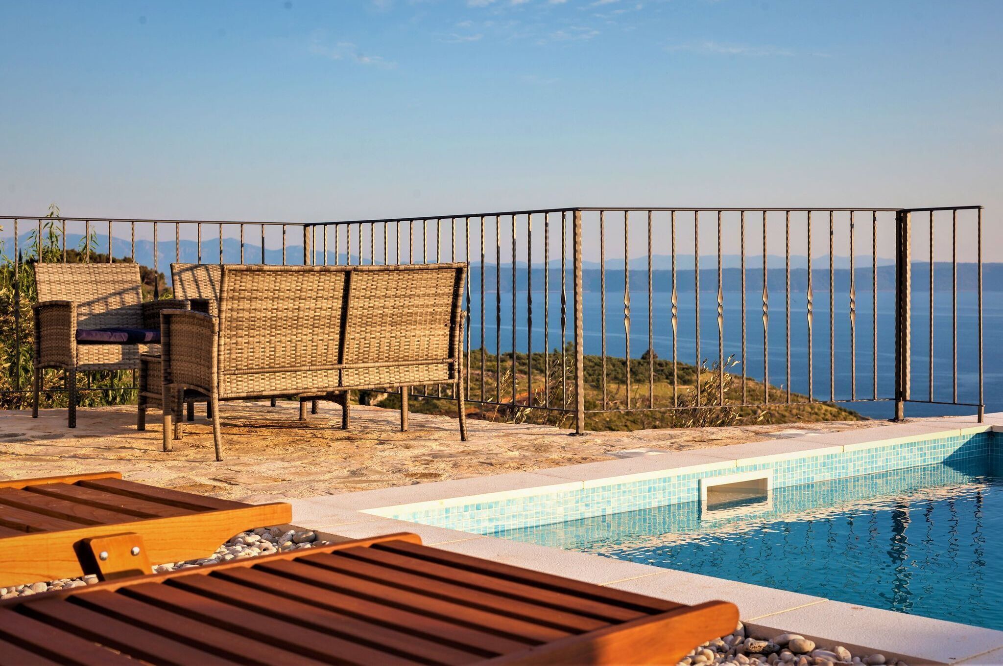 Ein Poolbereich mit Korbstühlen und einer Holzliege, einem Metallzaun und Blick auf das Meer.