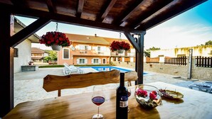 A wooden table with a bottle of wine, a glass, and a bowl of fruit in a covered outdoor area with a view of a swimming pool and buildings.