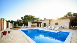 A swimming pool with white lounge chairs, a stone patio, and a small building with a pergola.
