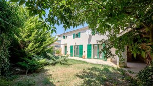 Un edificio blanco con contraventanas verdes y una antena parabólica, rodeado de vegetación y un sendero de piedra.