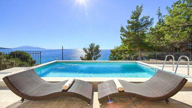 A rectangular swimming pool with two wicker lounge chairs and a metal ladder, surrounded by a metal fence, overlooking a body of water and trees. - Villa in Cres with Sea Views & Pool (Martinšćica)