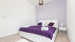 Ein Schlafzimmer mit einem weißen Bett, violetter Bettwäsche, einem weißen Nachttisch und einer weißen Tür.