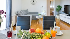 Una sala moderna con un sofá gris, dos sillas negras, una mesita blanca con una lámpara, un televisor sobre un mueble blanco y una cesta de fruta en la mesita.