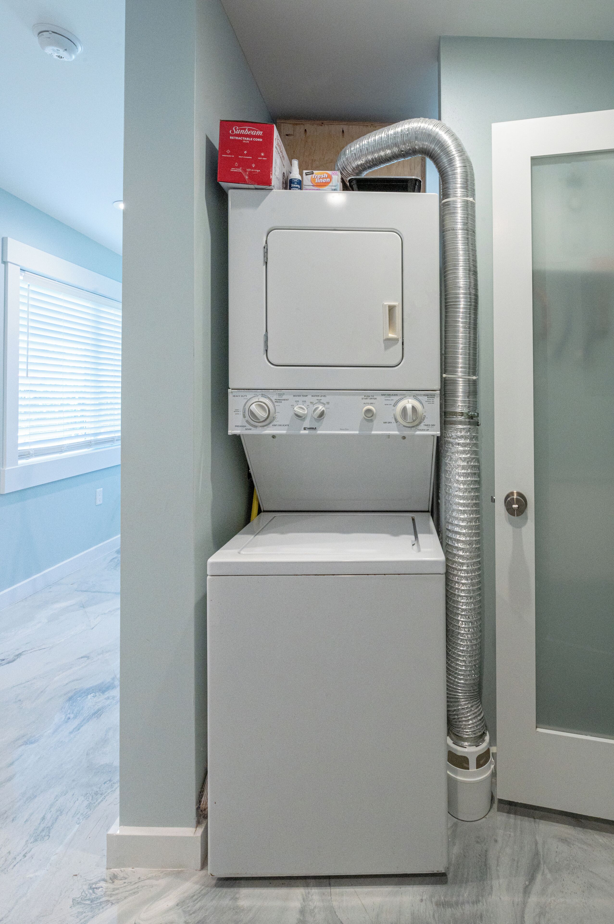 A compact, modern laundry room with a built-in washer and dryer, a vent hose, and a small shelf with cleaning supplies.