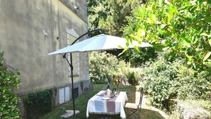 Un área de comedor al aire libre con una mesa bajo un paraguas a rayas, rodeada de follaje y un muro de edificio.