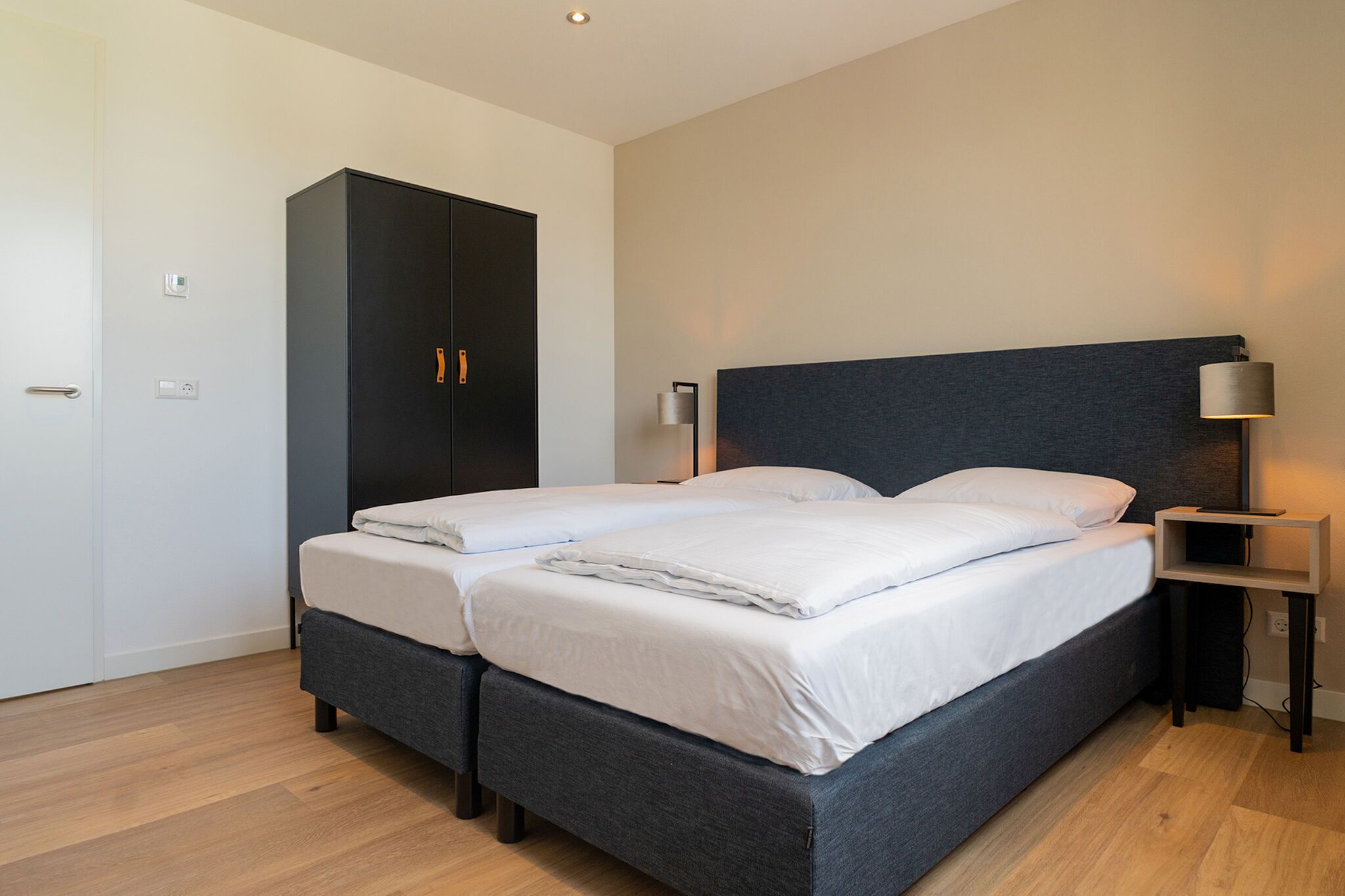 A double bed with a grey headboard, a wardrobe, and a bedside table with a lamp.