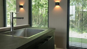 A modern bathroom with a large sink, frosted glass shower enclosure, and a view of greenery through the window blinds.