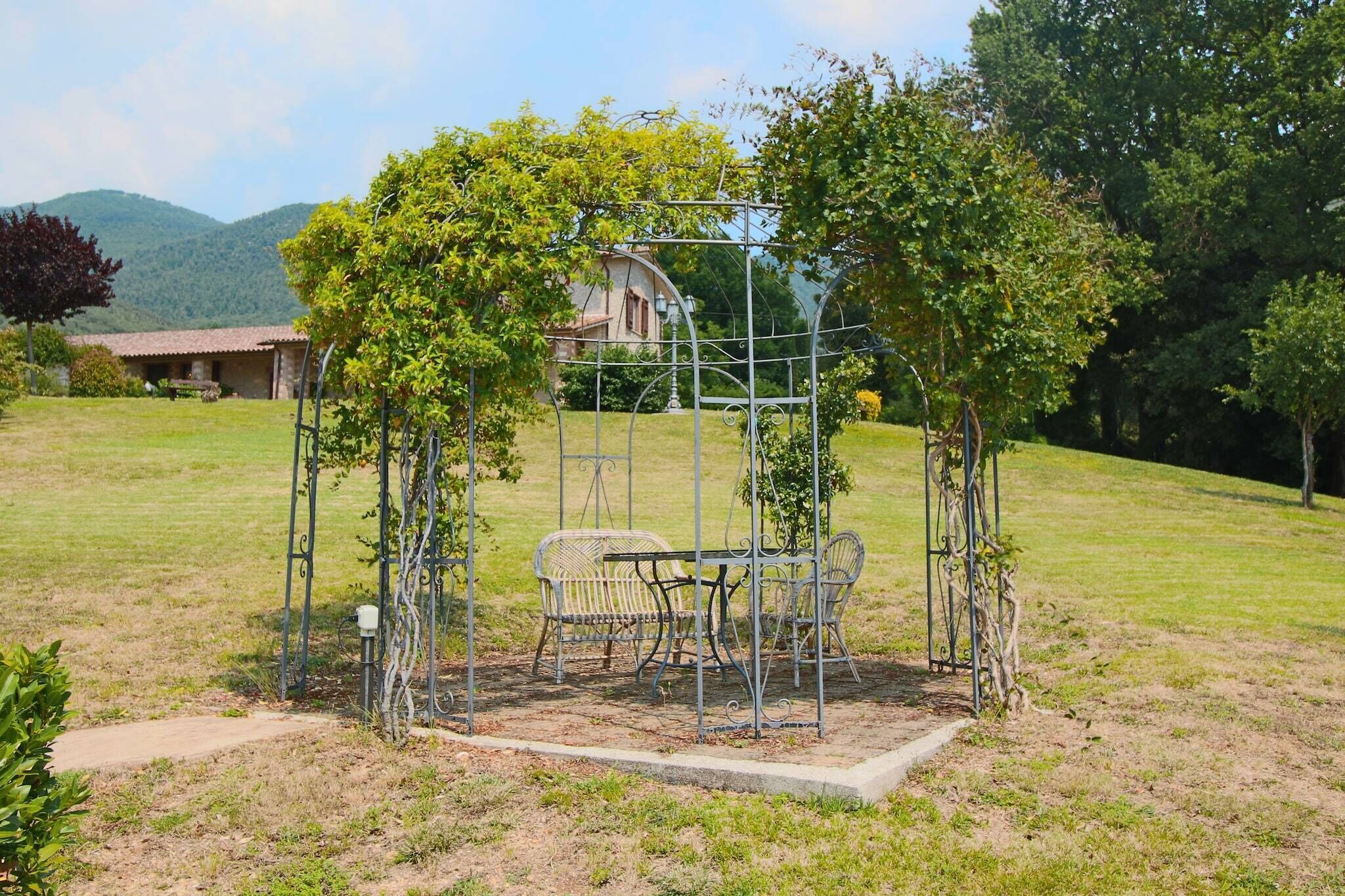 Um gazebo com cadeiras de metal e uma mesa em uma área gramada, com árvores e um edifício ao fundo.