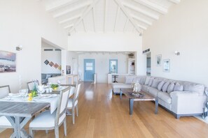 A spacious living room with a high ceiling, a dining area with a wooden table and chairs, and a large grey sofa.