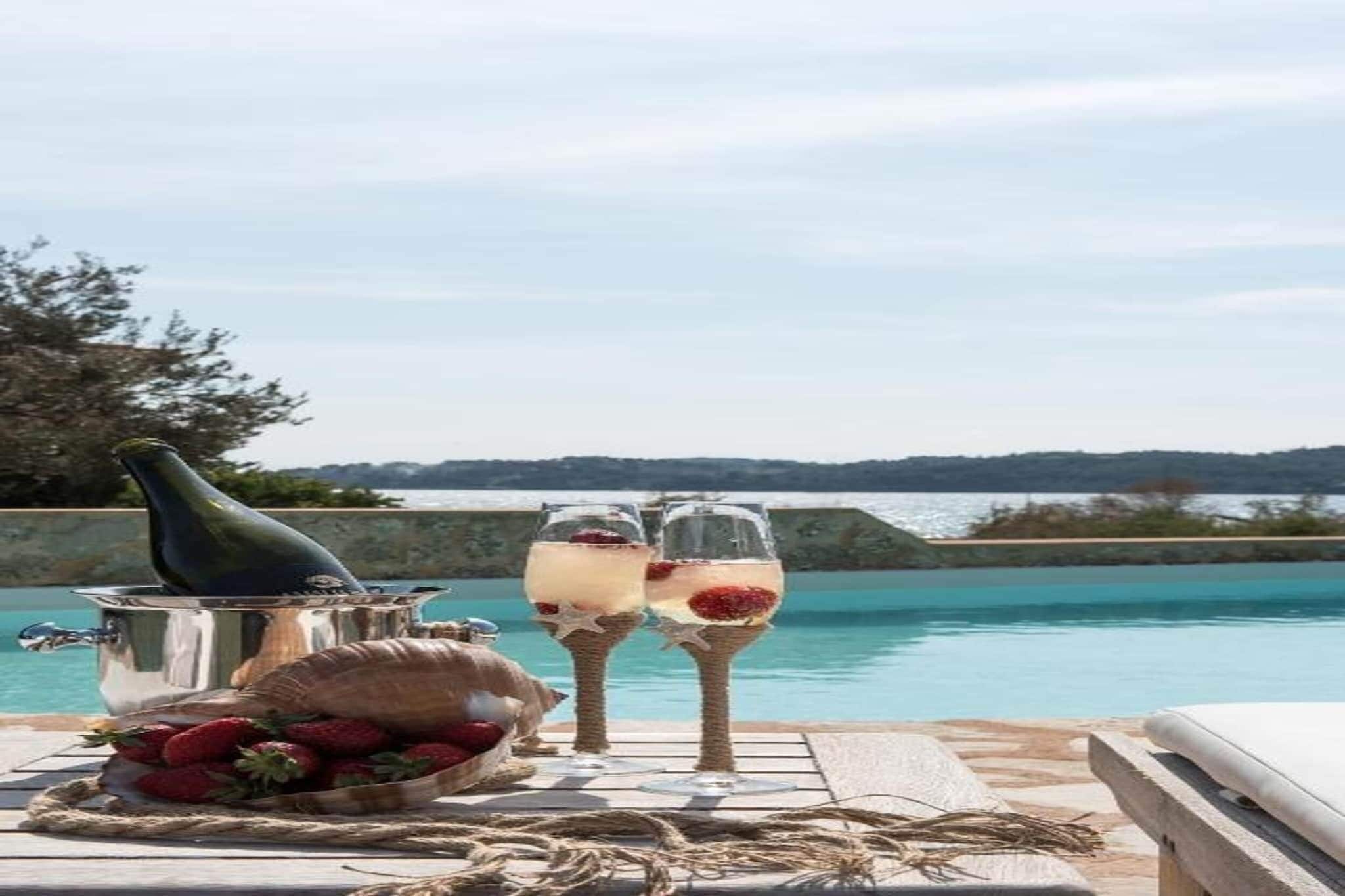 A table with a bottle, a basket of strawberries, and two glasses of a beverage with a red garnish, set against a backdrop of a body of water and distant hills.