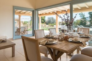 A dining area with a wooden table set for four, chairs, and a view of the outdoors through large glass doors. - Villa in Peloponnese with Beach & Pool (Porto Heli)