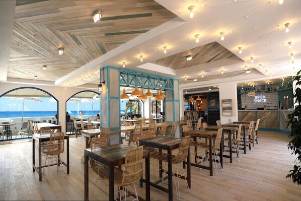 Ein Restaurant mit Holztischen und -stühlen, einem Barbereich und großen Fenstern mit Meerblick.