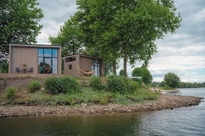 A modern building with large windows and a wooden exterior, situated by a river with a hammock and outdoor seating. - Tiny House by Dutch Waterfront Retreat (Maurik)