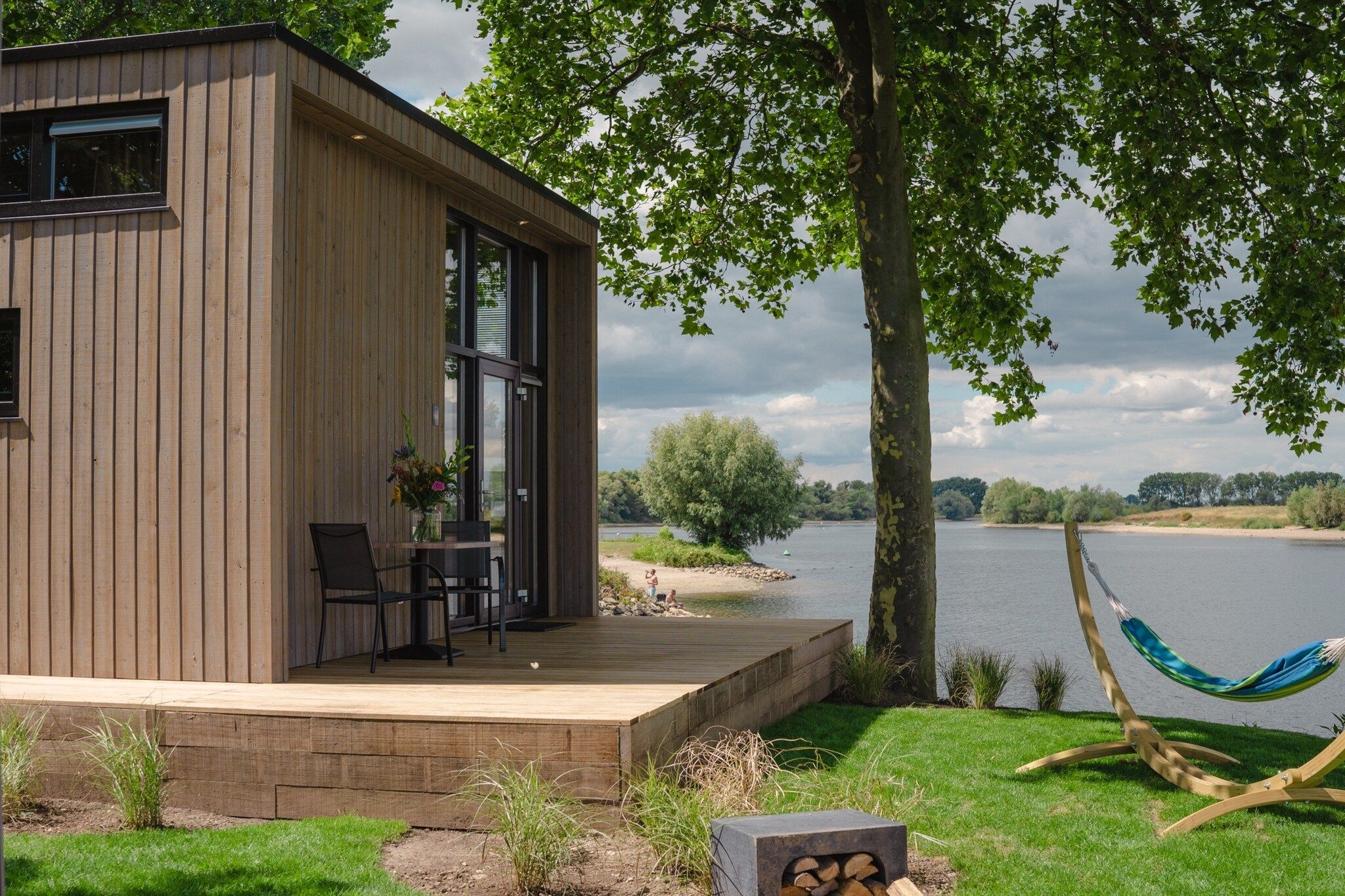 A modern wooden cabin with a deck, a hammock, and a view of a lake.