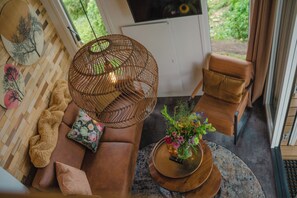 A cozy living room with a brown sofa, a woven floor lamp, a round coffee table with a floral arrangement, and a view of greenery outside. - Tiny House by Dutch Waterfront Retreat (Maurik)