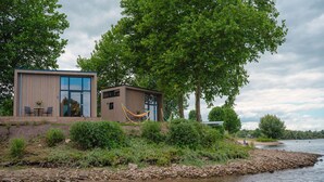 Una cabaña moderna junto a un lago, con comedor, hamaca y un cobertizo pequeño.