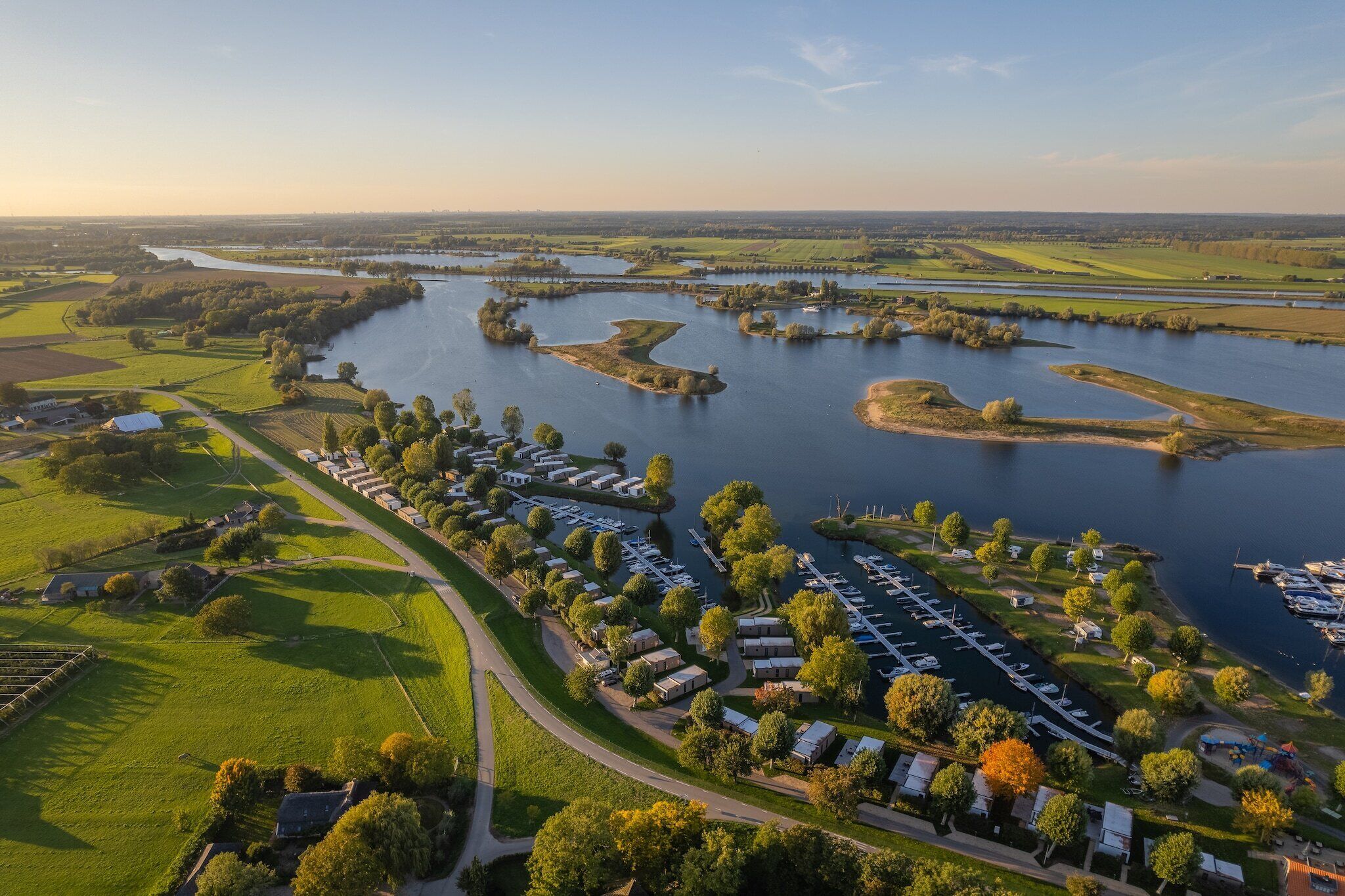 Vista aérea de un camping junto al río con varias áreas para vehículos recreativos, un puerto deportivo y campos verdes circundantes.