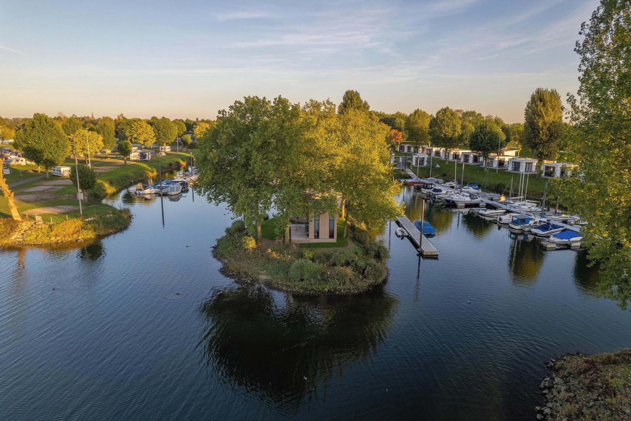 Una marina a la orilla del río con barcos amarrados, una pequeña isla con un edificio, y una hilera de casas móviles a lo largo de la ribera.