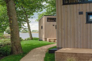 A wooden cabin with a gravel path leading to a grassy area and a body of water in the background. - Tiny House by Dutch Waterfront Retreat (Maurik)