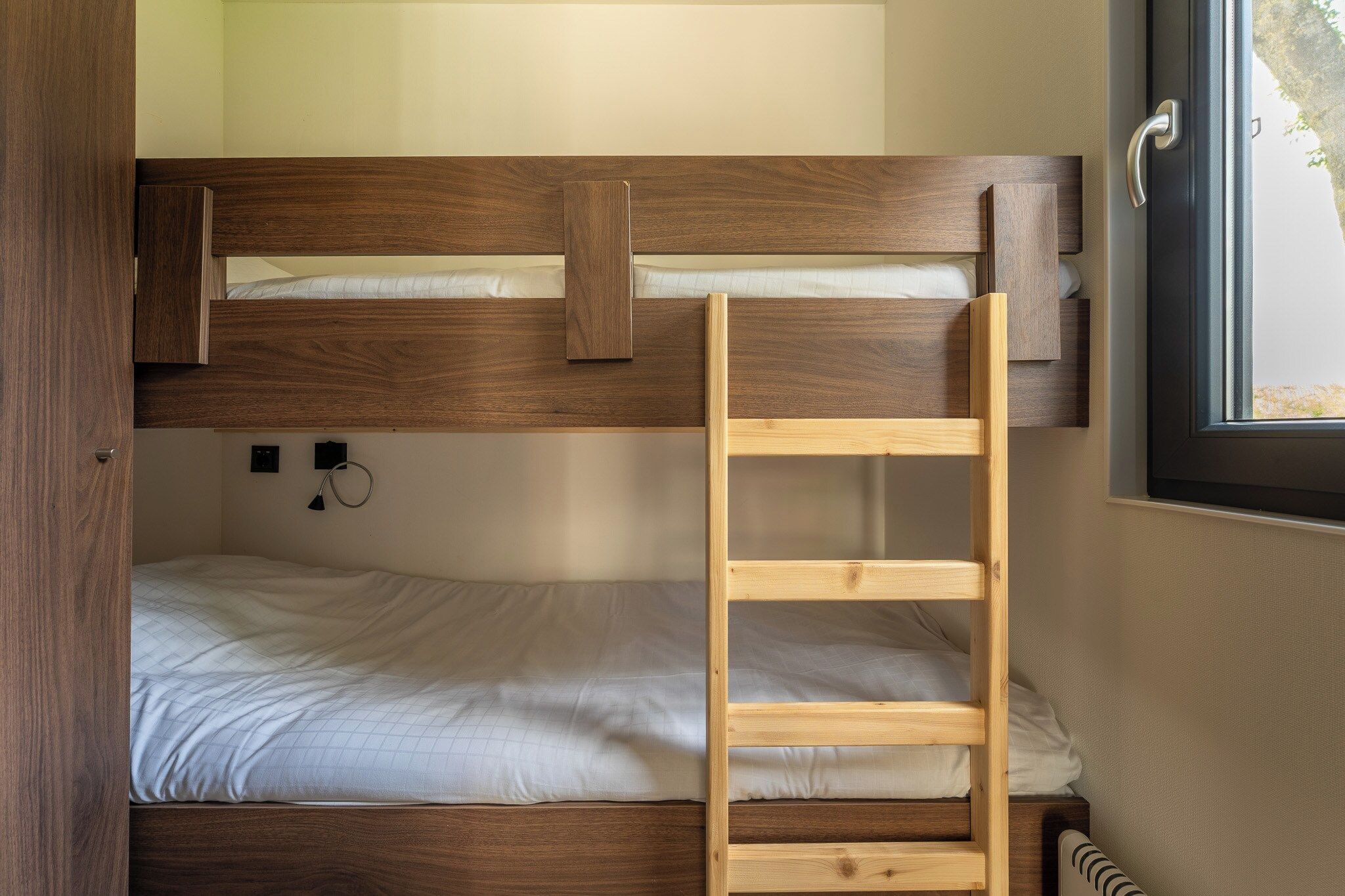 A bunk bed with a wooden ladder, a small desk with a chair, and a window with a view of greenery.