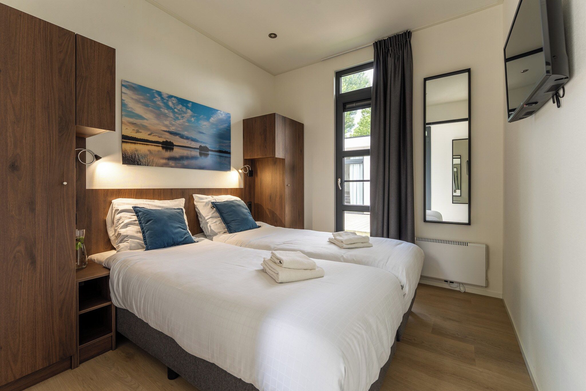 A double bed with white linens and blue pillows, a painting of a landscape, a window with curtains, and a wall-mounted mirror.