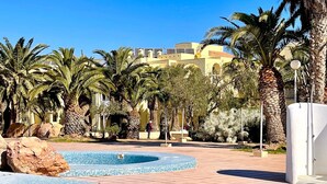 Zona de piscina con palmeras, un muro de piedra y un edificio al fondo.