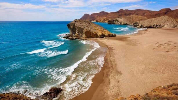 Un paisaje costero con una playa arenosa, afloramientos rocosos y montañas distantes bajo un cielo despejado.