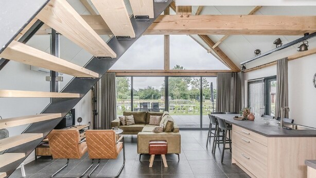 A modern interior with a wooden staircase, a sofa, a dining area, and large windows offering a view of greenery.