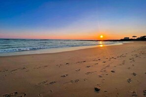 A beach with footprints leading to the water, a sunset in the background, and a building on the right.