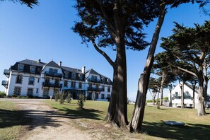 Um edifício grande, com vários andares, fachada simétrica, entrada central e um gramado bem cuidado.