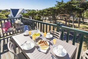 Mesa de jantar posta com pratos, talheres e um jarro de suco em varanda com vista para árvores e um edifício.