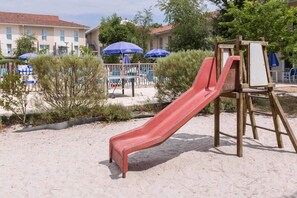 A sandy area with a red slide and an easel with a blank canvas, surrounded by greenery and buildings in the background.