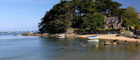 A coastal scene with a sandy beach, calm water, boats, and a stone building with a large tree.