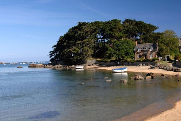 A coastal scene with a sandy beach, calm water, boats, and a stone building with a large tree.