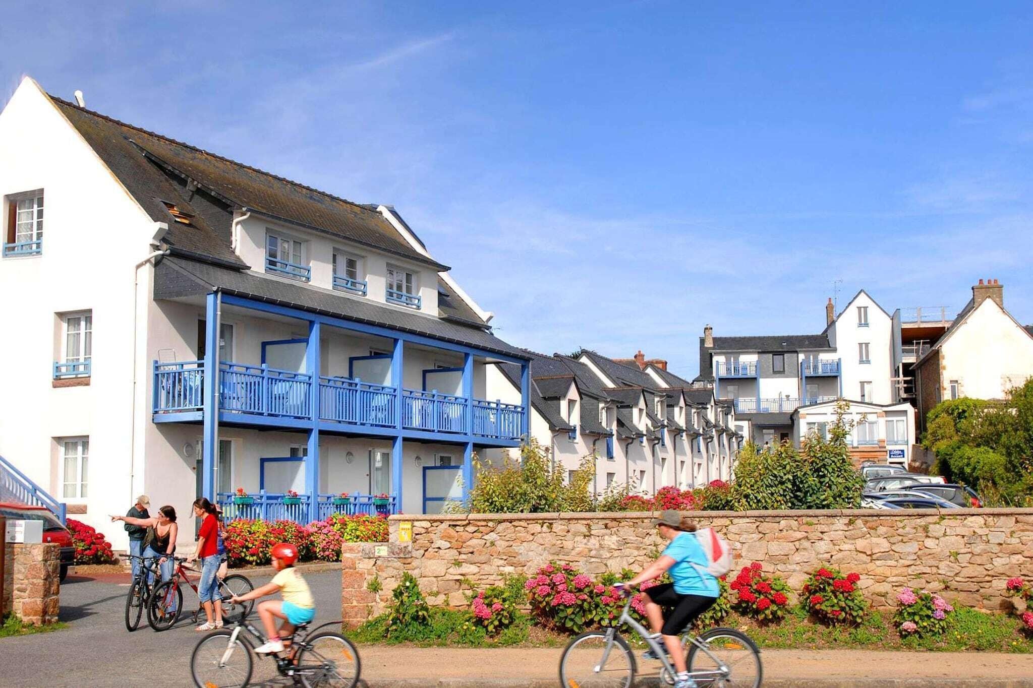 A row of white buildings with blue balconies, a stone wall with flowers, and people riding bicycles.