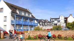 Weiße Häuser mit blauen Balkonen, eine Steinmauer mit Blumen und Radfahrer.