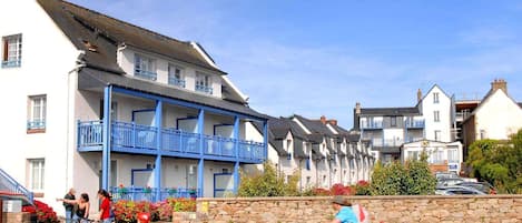 A row of white buildings with blue balconies, a stone wall with flowers, and people riding bicycles.