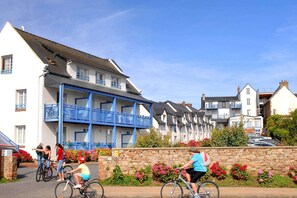 Weiße Häuser mit blauen Balkonen, eine Steinmauer mit Blumen und Radfahrer.