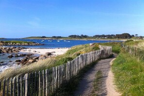 Ein Küstenpfad mit Holzzaun, der zu einem Strand mit Booten und blauem Himmel führt.
