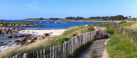 A coastal path with a wooden fence, leading to a beach with boats and a clear sky.