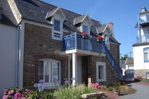 A two-story building with a stone facade, a blue balcony with flower boxes, and a small staircase leading to the upper floor.