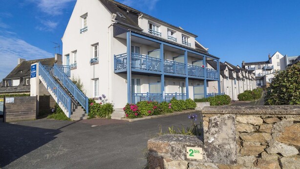 A two-story building with a blue staircase and balcony railings, a stone wall with a number plaque, and a clear blue sky. - Apartment in Trégastel near the Beach (Trégastel)