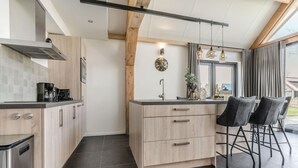 A modern kitchen with wooden cabinets, a stainless steel range hood, and a bar area with two black bar stools. - Holiday Home in Netherlands Pond View (Baarle-Nassau)