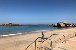 A beach with clear skies, a person standing near a metal structure, and large rocks in the water.