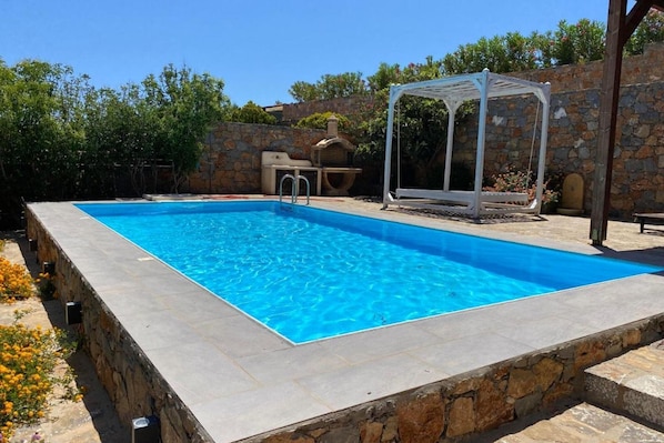 A rectangular swimming pool with clear blue water, surrounded by stone walls and a pergola structure. - Villa in Elounda with Private Pool & BBQ (Elounda)