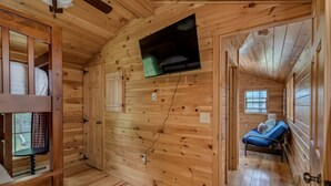 Interior de uma cabana de madeira com uma cama, uma televisão fixada na parede e uma janela.